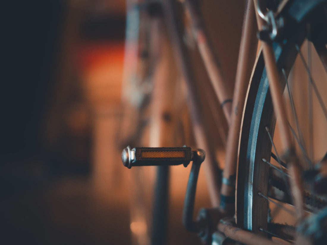Pedal eines Alltagsrads mit orangefarbenem Reflektor im Abendlicht, dahinter Kettenstrebe und Hinterrad