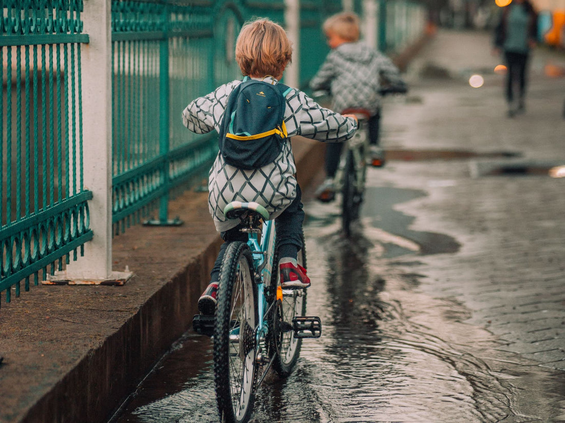 Zwei Kinder fahren auf einer nassen Straße hintereinander Rad, Wasser und Schmutz spritzen an Kette und Reifen