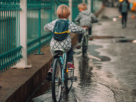 Zwei Kinder fahren auf einer nassen Straße hintereinander Rad, Wasser und Schmutz spritzen an Kette und Reifen
