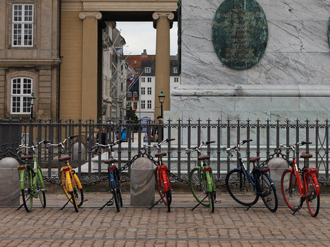 Sieben bunte Stadträder stehen nebeneinander auf ihren Hinterbauständern an einem Geländer auf einem Kopfsteinpflasterplatz