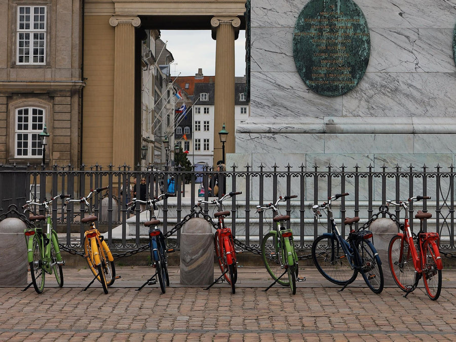 Sieben bunte Stadträder stehen nebeneinander auf ihren Hinterbauständern an einem Geländer auf einem Kopfsteinpflasterplatz