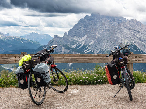Zwei bepackte Tourenräder mit Packtaschen vorn und hinten stehen an einem Holzgeländer vor einem Bergpanorama