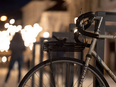 Frontgepäckträger über dem Vorderrad eines silbernen Rennrads, das Rad steht abends an einem Fahrradbügel in einer beleuchteten Straße