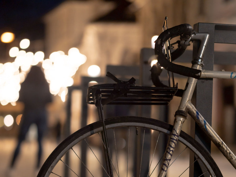 Frontgepäckträger über dem Vorderrad eines silbernen Rennrads, das Rad steht abends an einem Fahrradbügel in einer beleuchteten Straße